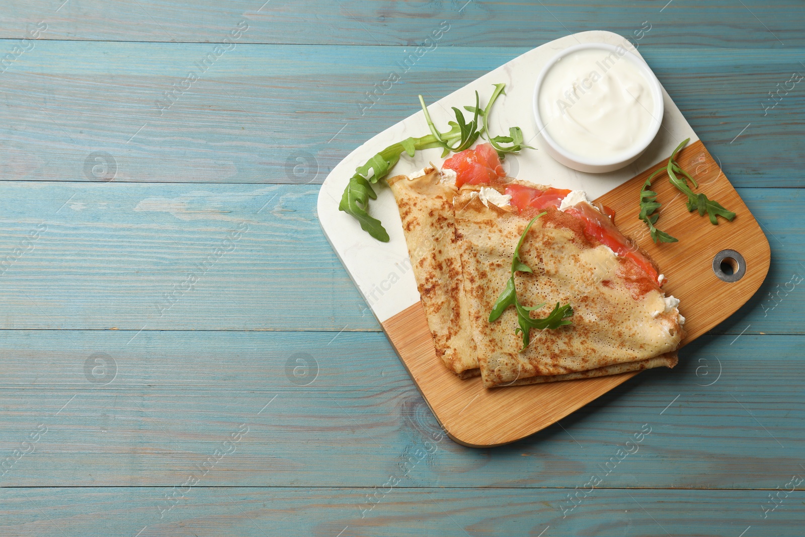 Photo of Delicious crepes with salmon, cream cheese and arugula on blue wooden table, top view. Space for text