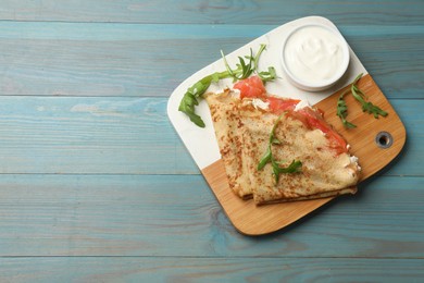 Photo of Delicious crepes with salmon, cream cheese and arugula on blue wooden table, top view. Space for text