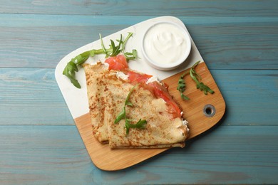 Photo of Delicious crepes with salmon, cream cheese and arugula on blue wooden table, top view