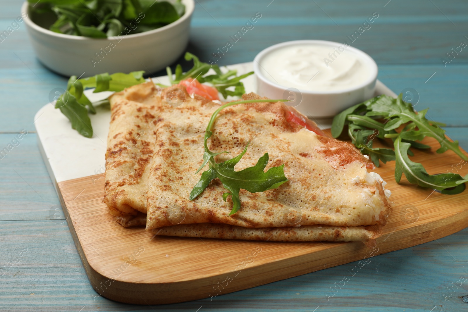 Photo of Delicious crepes with salmon, cream cheese and arugula on blue wooden table, closeup