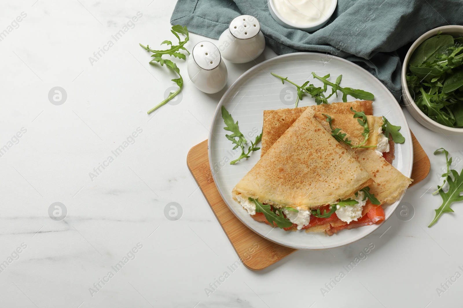 Photo of Delicious crepes with salmon, cream cheese and arugula on white marble table, flat lay. Space for text