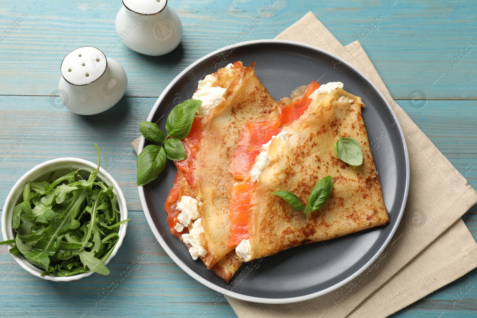 Photo of Delicious crepes with salmon, cream cheese, basil and arugula on light blue wooden table, flat lay