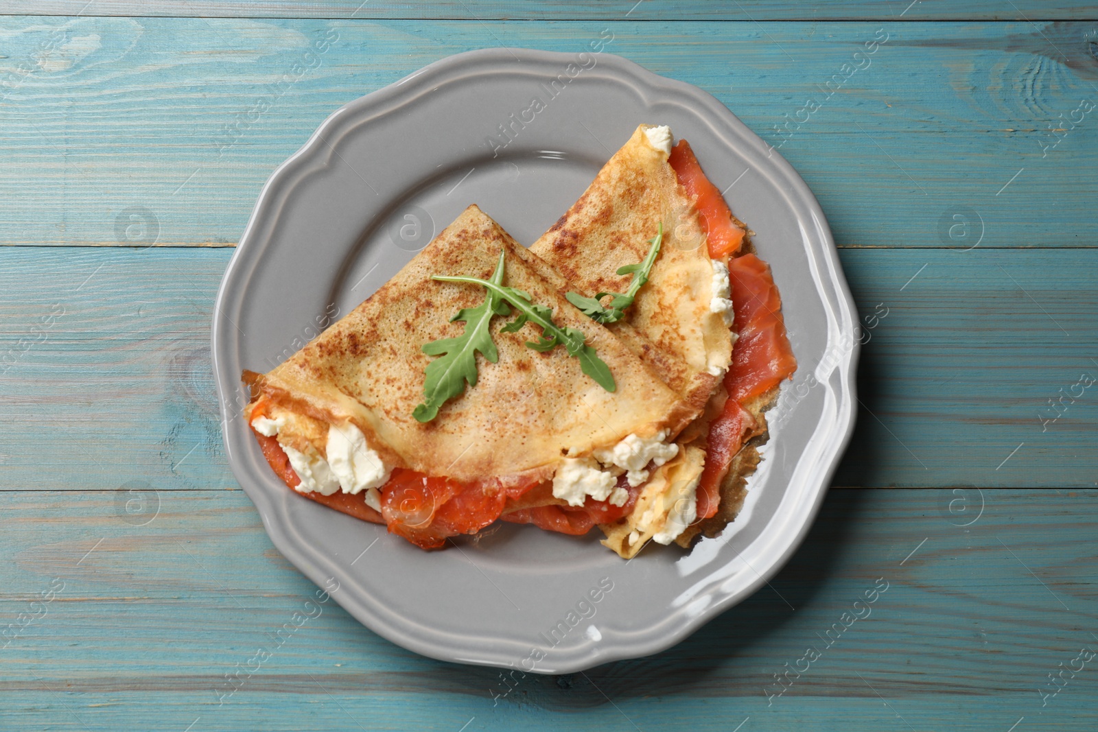 Photo of Delicious crepes with salmon, cream cheese and arugula on light blue wooden table, top view