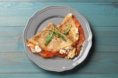 Photo of Delicious crepes with salmon, cream cheese and arugula on light blue wooden table, top view