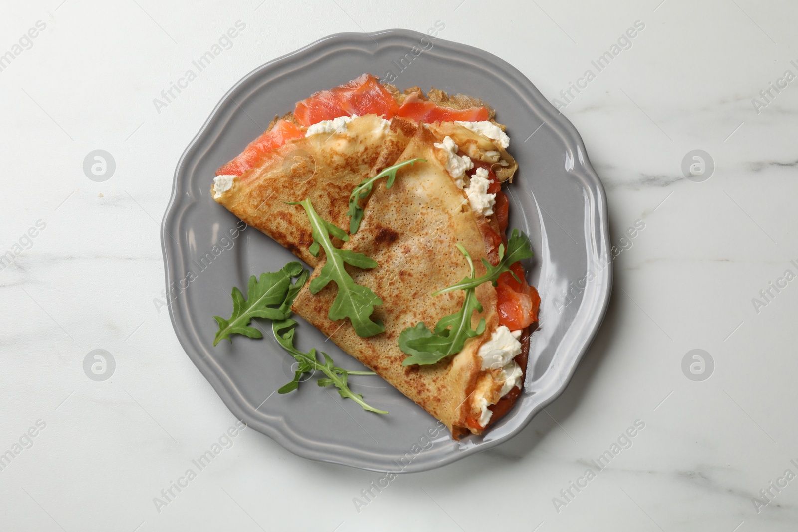 Photo of Delicious crepes with salmon, cream cheese and arugula on white marble table, top view