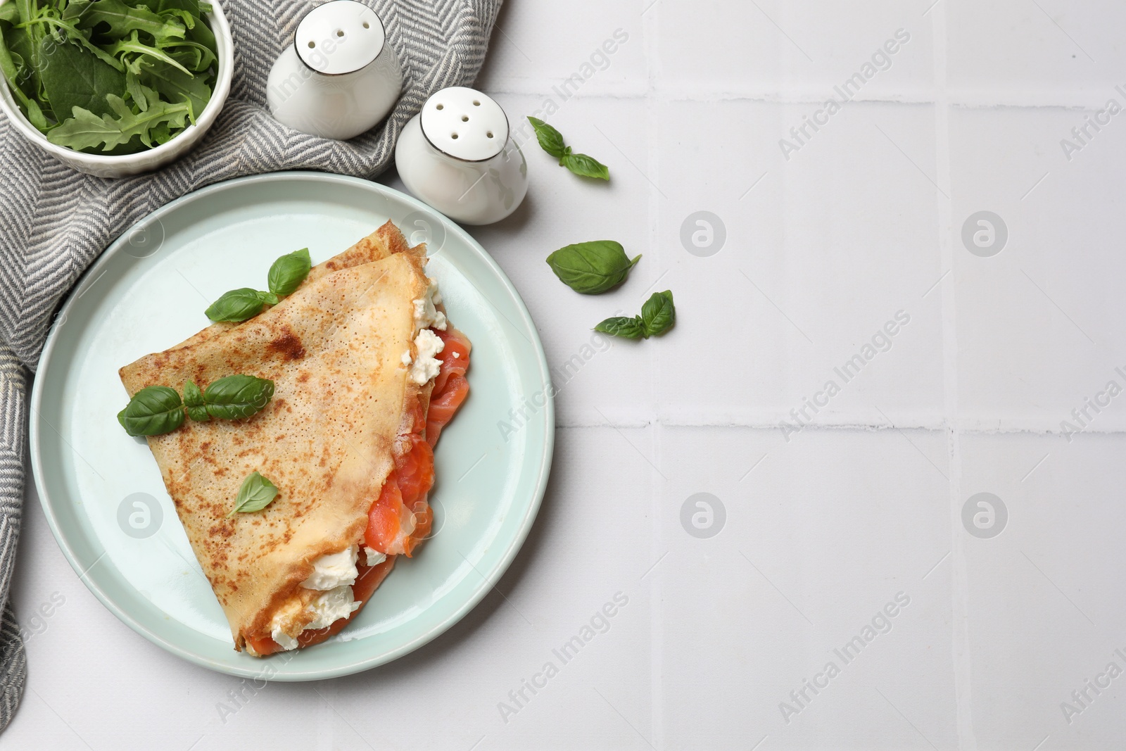 Photo of Delicious crepe with salmon, cream cheese, basil and arugula on white tiled table, flat lay. Space for text