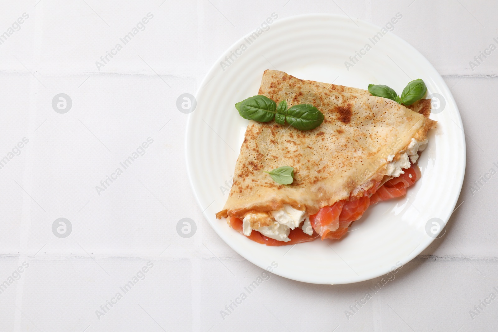 Photo of Delicious crepe with salmon, cream cheese and basil on white tiled table, top view. Space for text