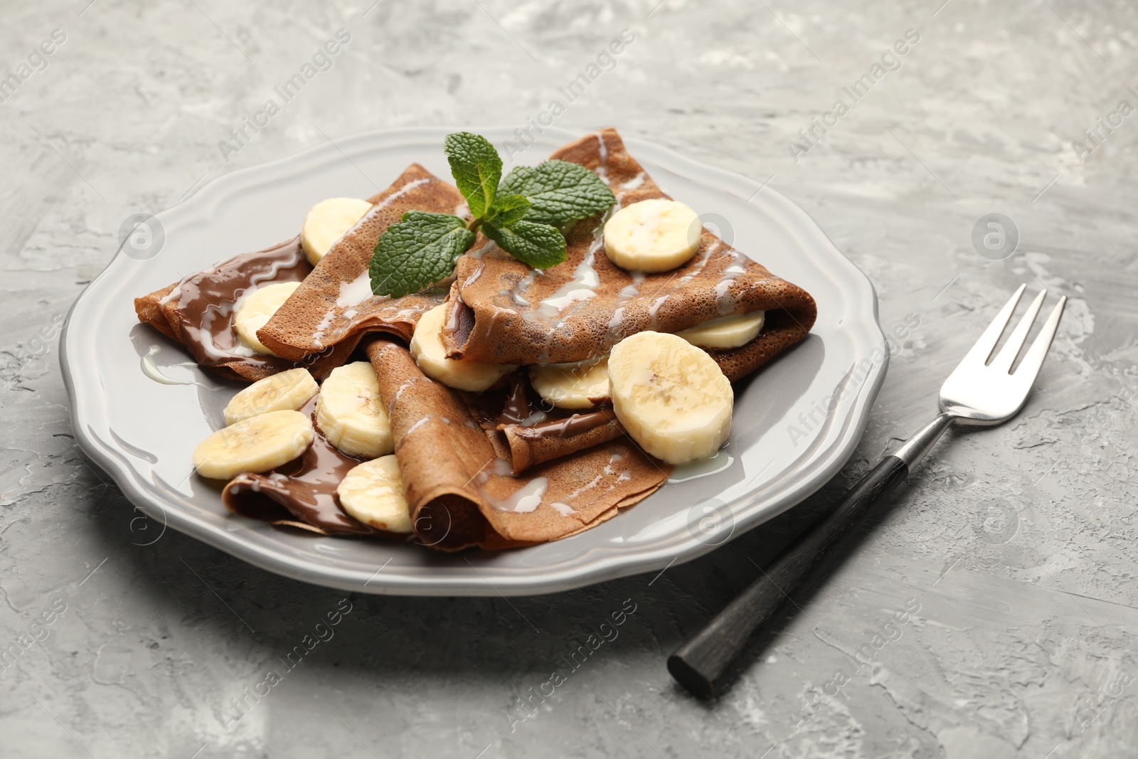 Photo of Delicious chocolate crepes with banana, condensed milk and mint served on grey table, closeup