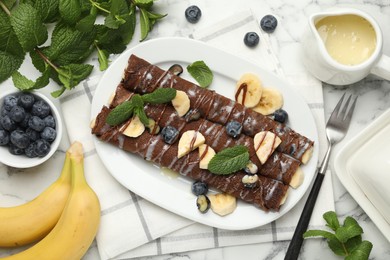 Photo of Delicious chocolate crepes with banana and blueberries served on white marble table, flat lay