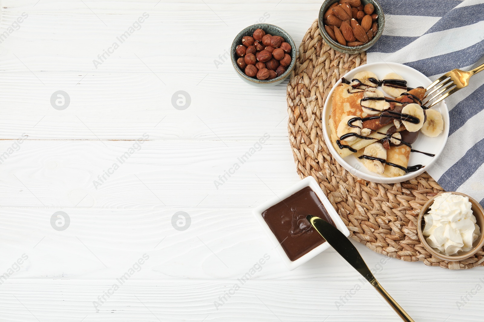 Photo of Delicious crepes with banana, chocolate sauce, nuts and whipped cream on white wooden table, flat lay. Space for text