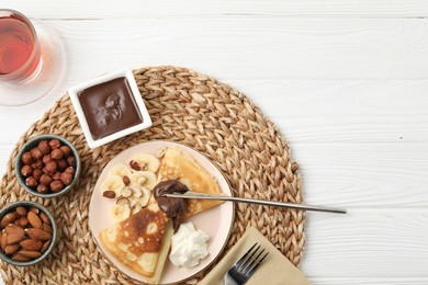 Photo of Delicious crepes with banana, chocolate paste, whipped cream, nuts and tea on white wooden table, flat lay