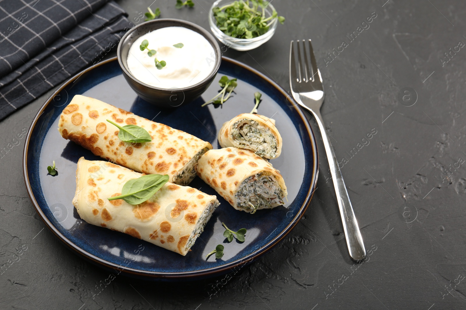 Photo of Delicious crepes with cottage cheese, spinach and sour cream served on black table, closeup