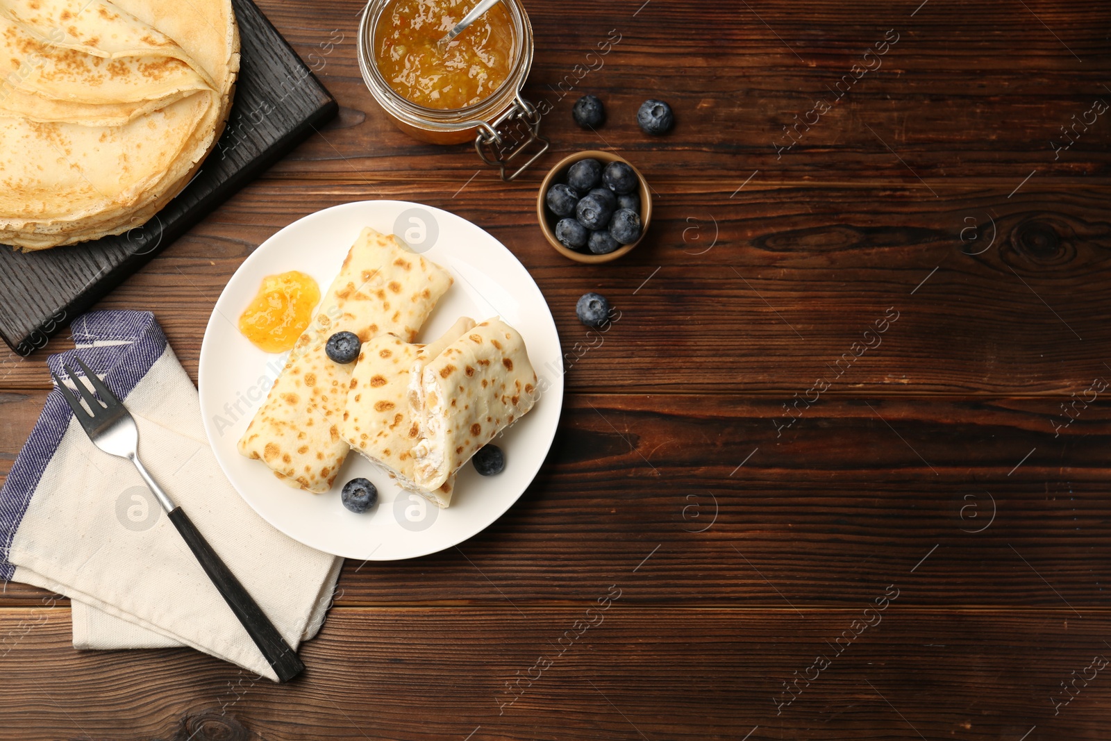 Photo of Delicious crepes with cottage cheese, jam and blueberries served on wooden table, flat lay. Space for text