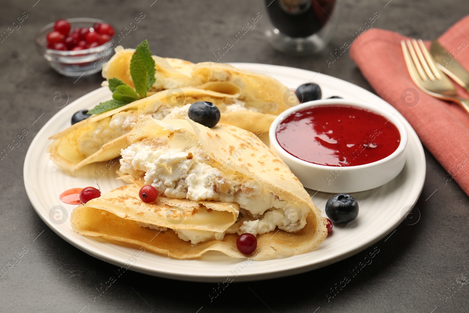 Photo of Delicious crepes with cottage cheese, jam, redcurrants and blueberries served on grey table, closeup