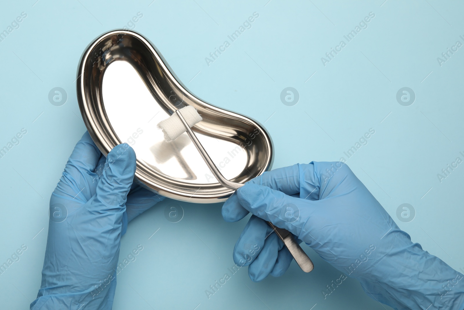 Photo of Doctor holding surgical tweezers with gauze and tray on light blue background, top view