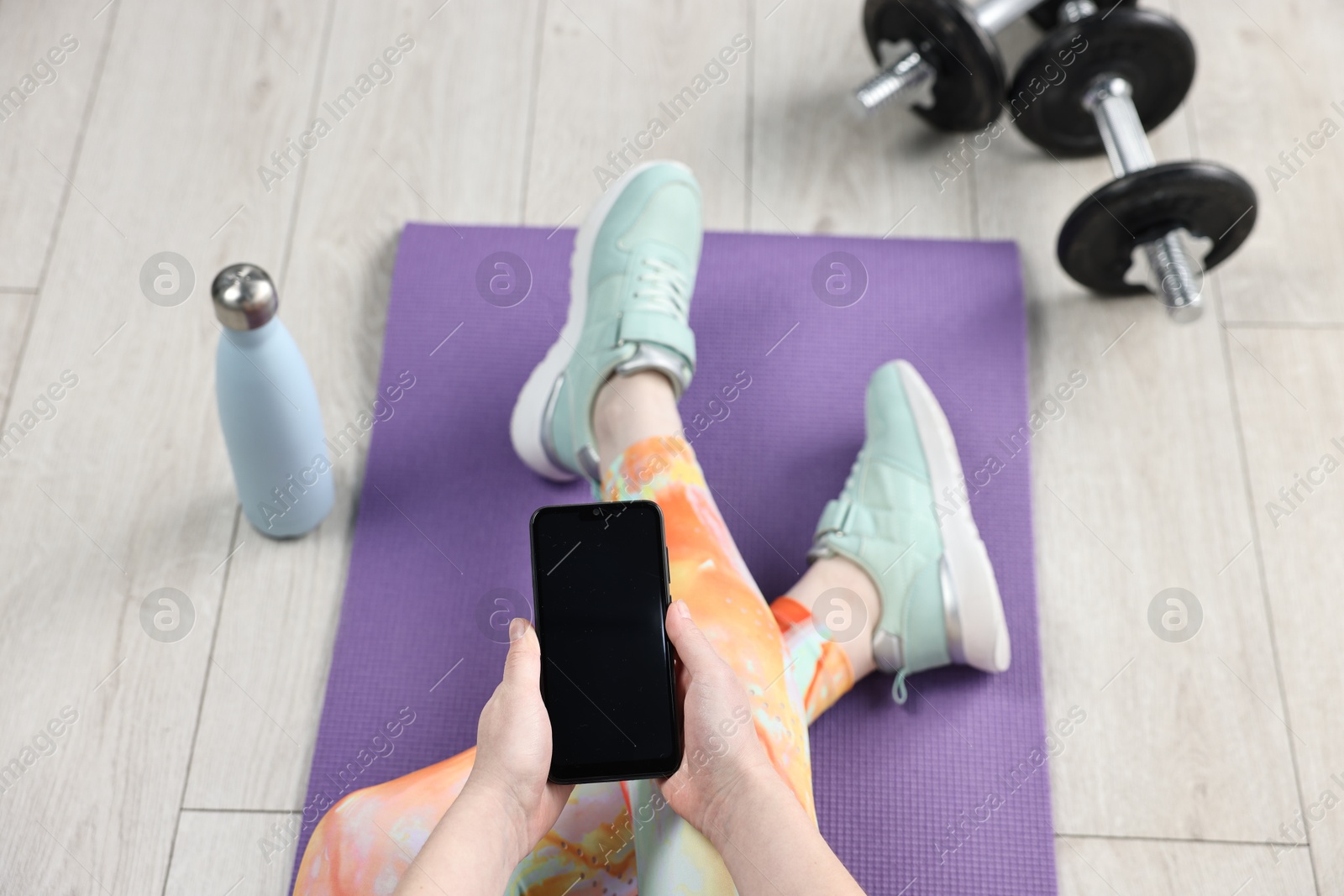 Photo of Woman with smartphone and gym equipment on floor indoors, above view