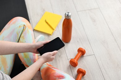 Photo of Woman with smartphone, gym equipment and planner on floor indoors, closeup. Space for text