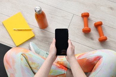 Photo of Woman with smartphone, gym equipment and planner on floor indoors, above view