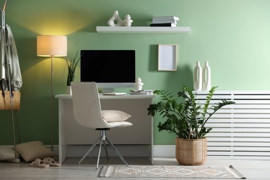 Photo of Home office. Comfortable workplace with desk, computer and lamp near light green wall in room