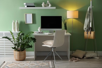 Photo of Home office. Comfortable workplace with desk, computer and lamp near light green wall in room