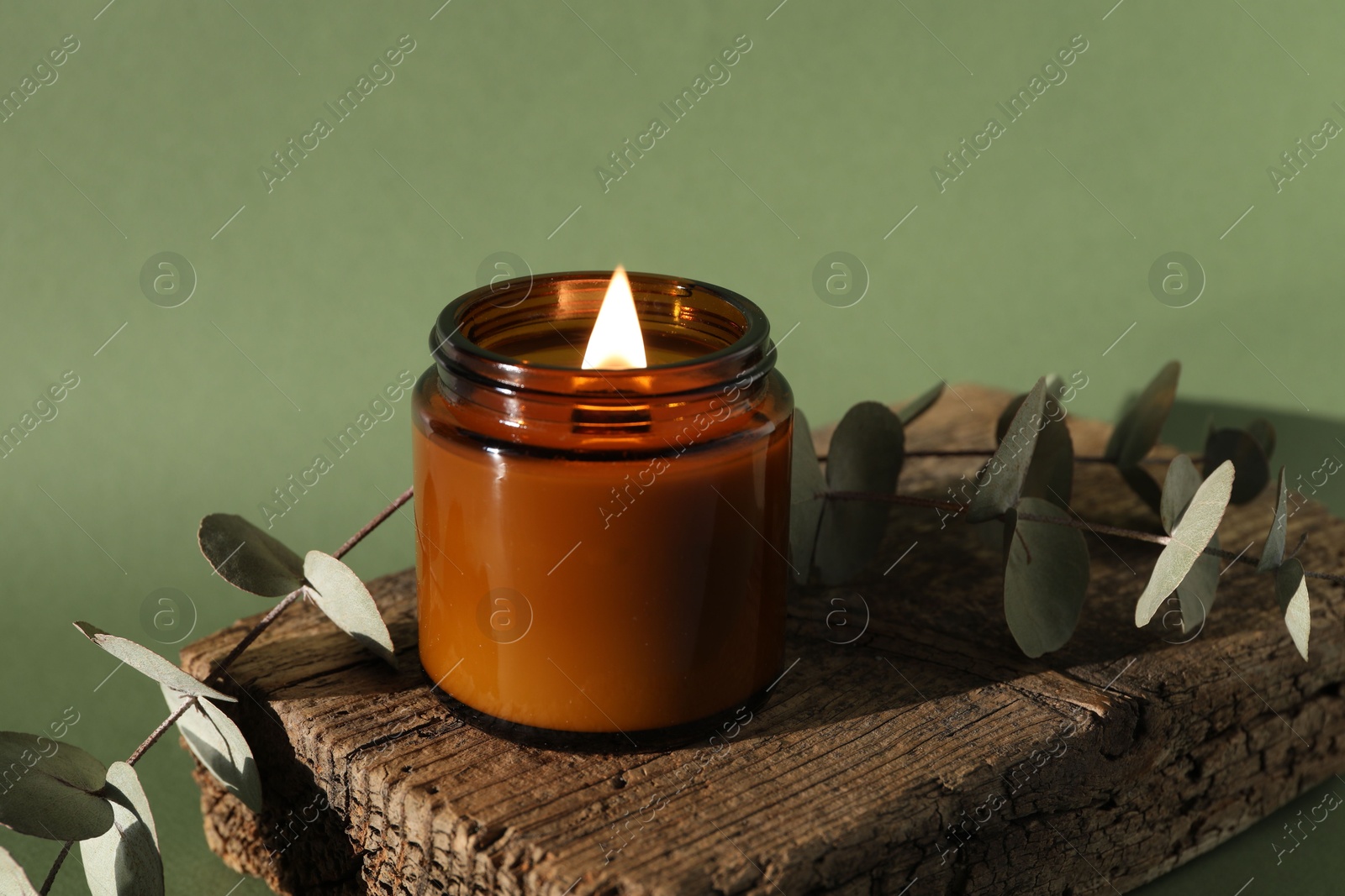 Photo of Burning soy wax candle, decorative piece of wood and eucalyptus branches on green background