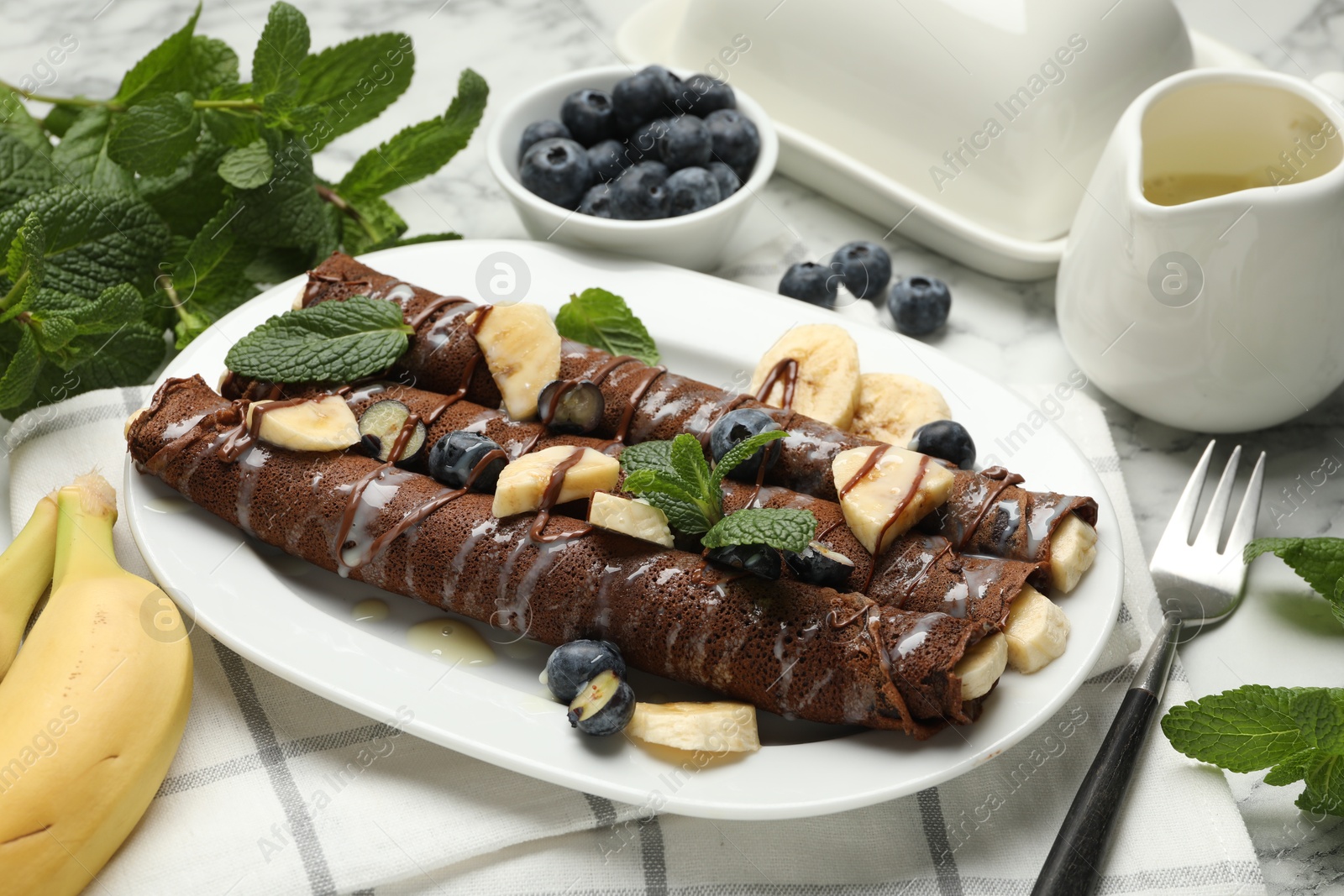 Photo of Delicious chocolate crepes with banana and blueberries served on white marble table, closeup