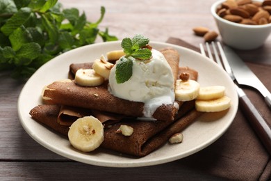 Photo of Delicious chocolate crepes with banana, nuts, mint and scoop of ice cream on wooden table, closeup