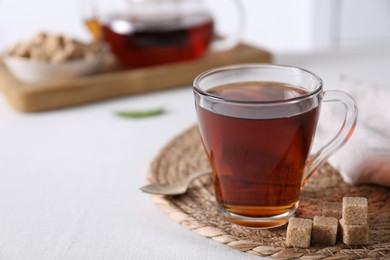 Photo of Aromatic black tea in cup and brown sugar cubes on white table, closeup. Space for text