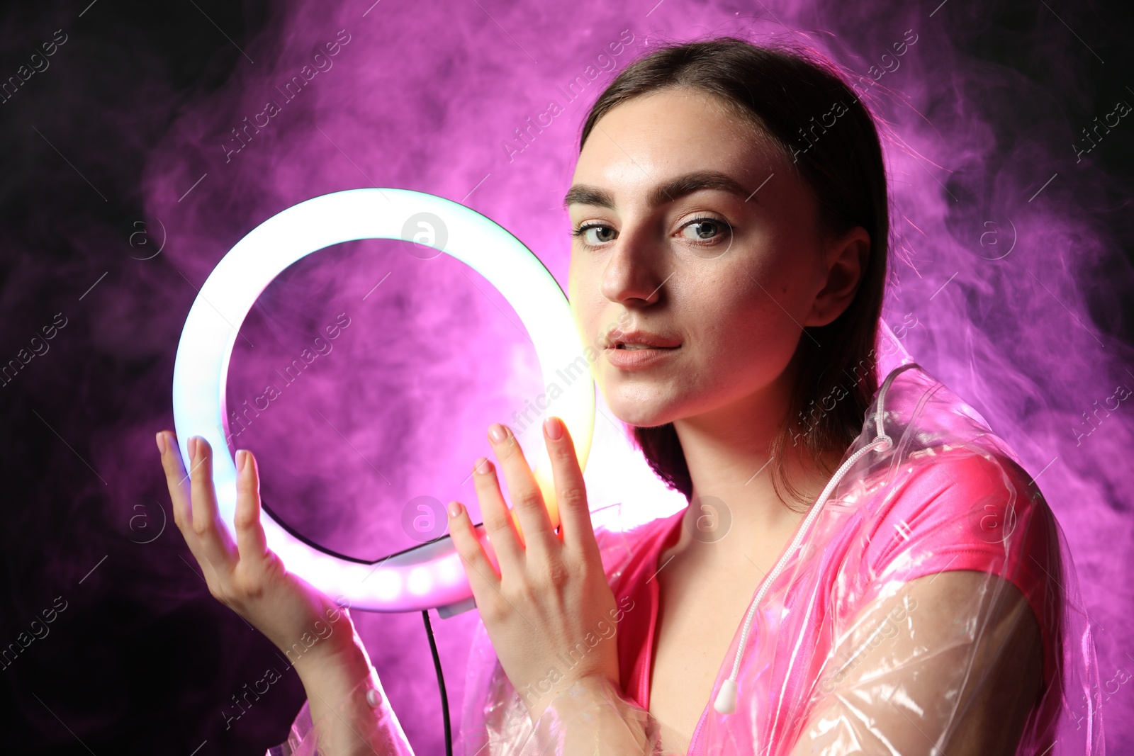 Photo of Beautiful woman with ring lamp wearing transparent coat on dark purple background in smoke