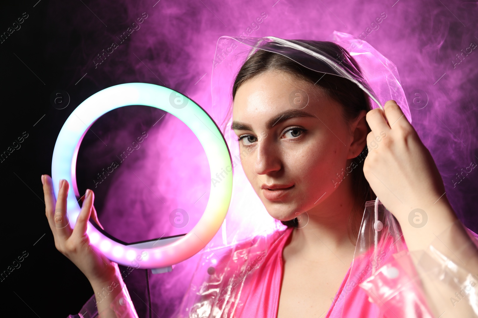 Photo of Beautiful woman with ring lamp wearing transparent coat on dark purple background in smoke