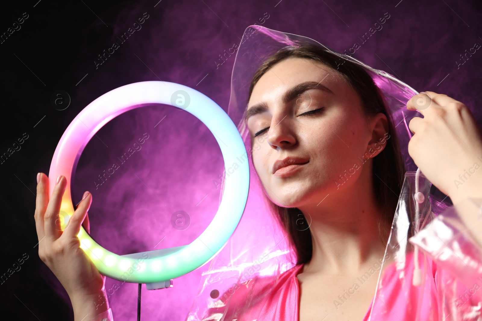 Photo of Beautiful woman with ring lamp wearing transparent coat on dark purple background in smoke, low angle view