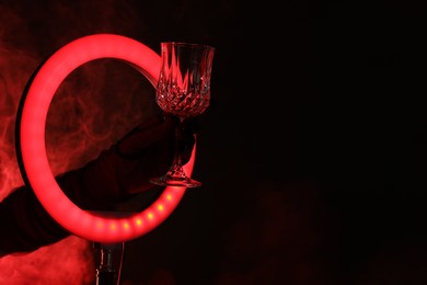 Photo of Woman holding empty wineglass through ring lamp on dark red background in smoke, closeup. Space for text