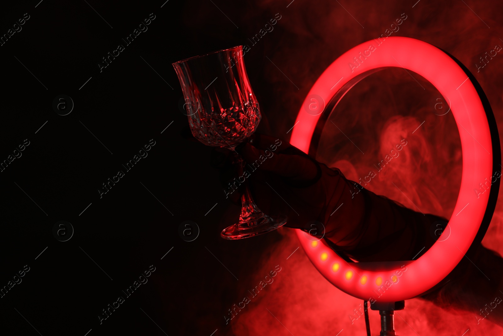 Photo of Woman holding empty wineglass through ring lamp on dark red background in smoke, closeup. Space for text