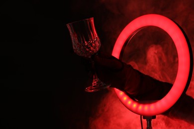 Photo of Woman holding empty wineglass through ring lamp on dark red background in smoke, closeup. Space for text