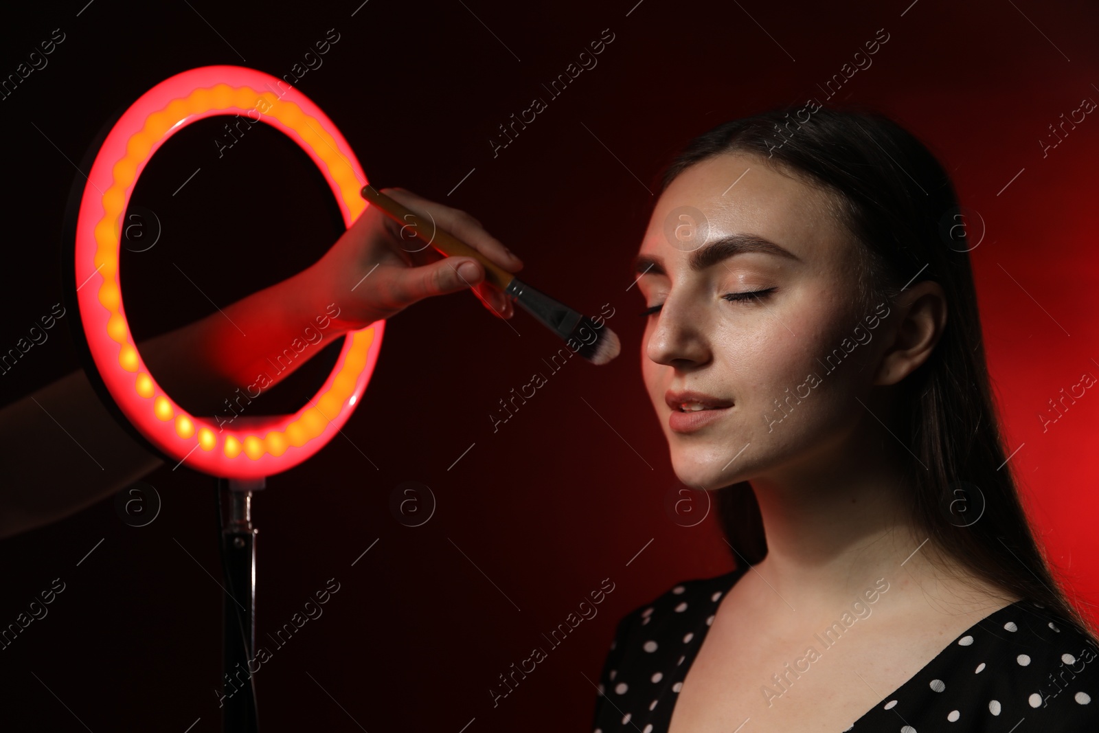 Photo of Makeup artist working with beautiful woman on dark red background, closeup. Using ring lamp