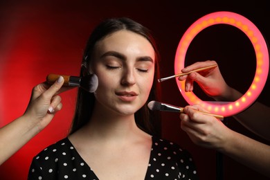 Photo of Makeup artists working with beautiful woman on dark red background, closeup. Using ring lamp