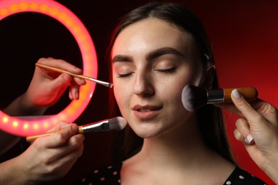 Photo of Makeup artists working with beautiful woman on dark red background, closeup. Using ring lamp