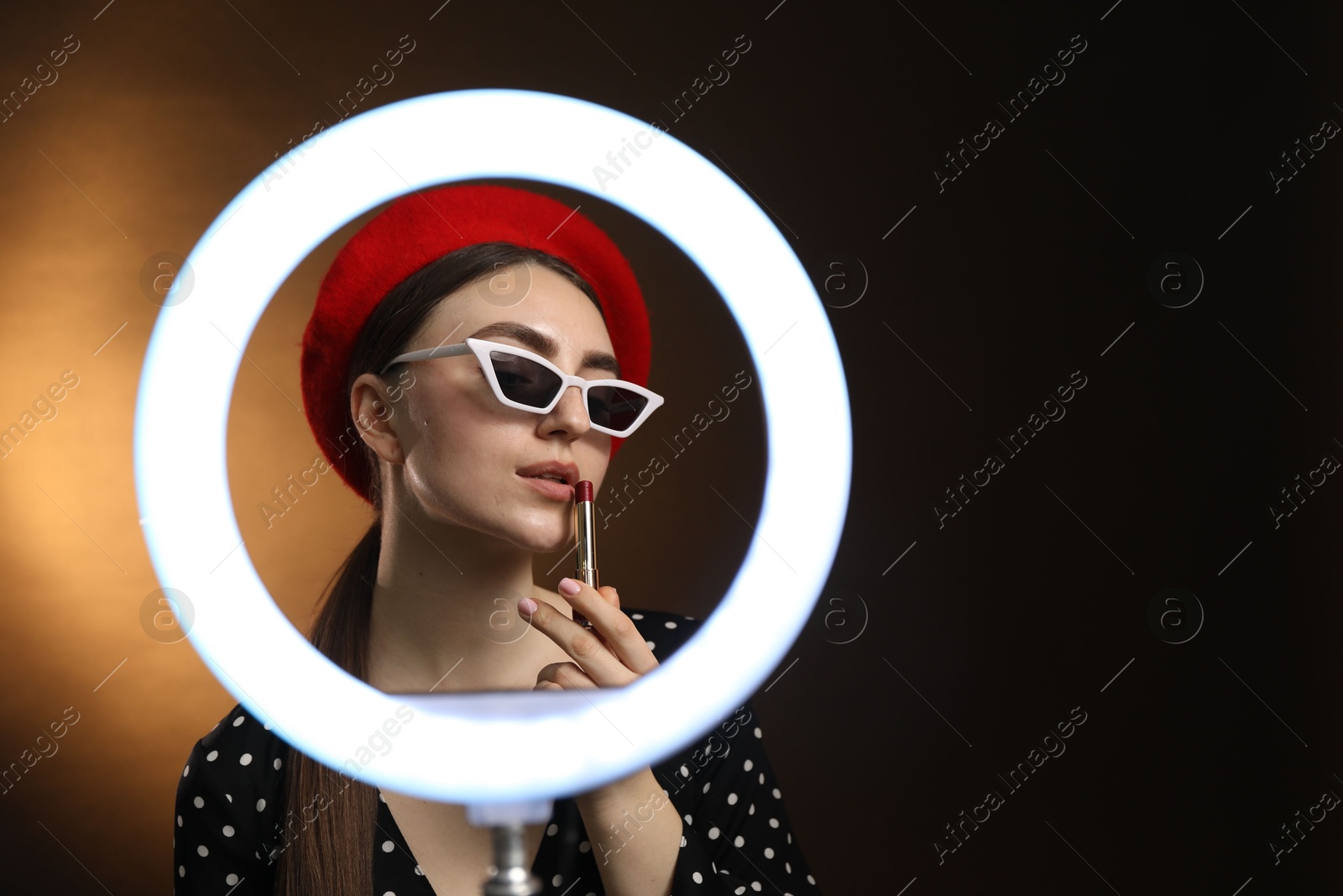 Photo of Beautiful woman applying lipstick on dark background, view through ring lamp. Space for text