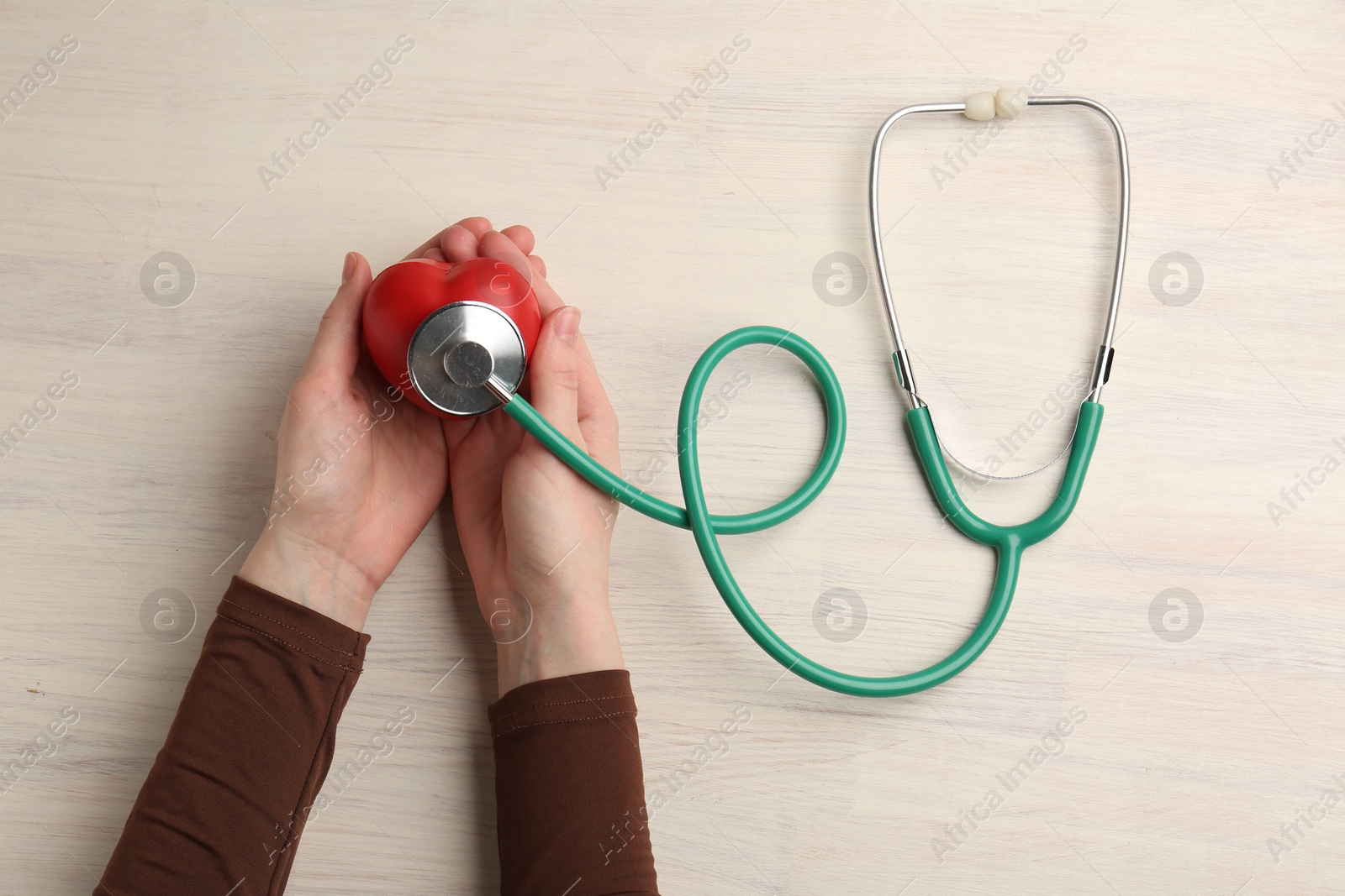 Photo of Cardiology. Woman with stethoscope and red decorative heart on light wooden table, top view