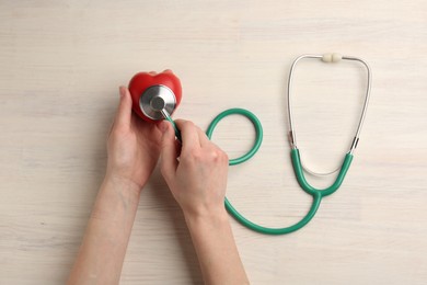 Photo of Cardiology. Woman with stethoscope and red decorative heart on light wooden table, top view