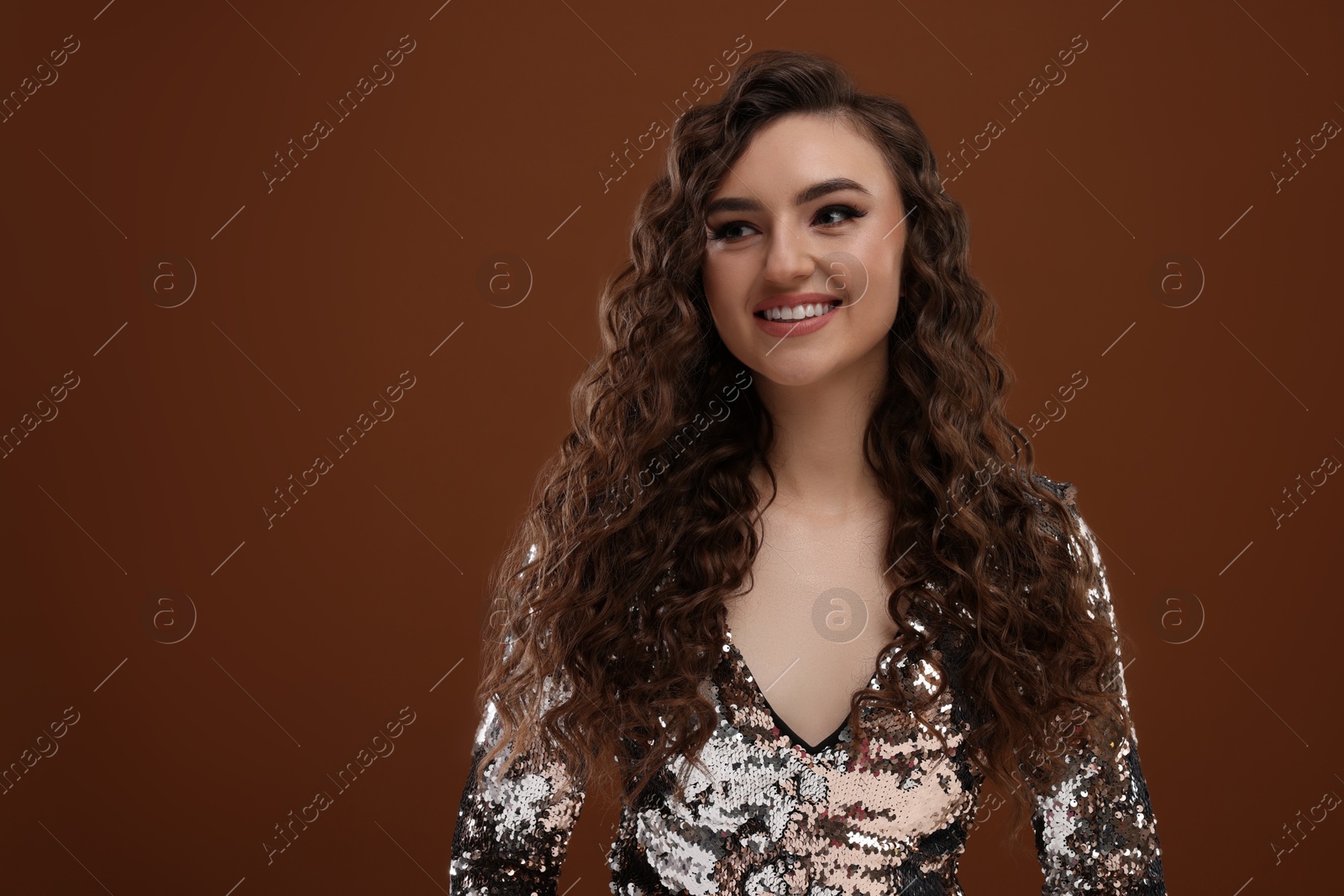 Photo of Beautiful young woman with long curly hair in sequin dress on brown background, space for text