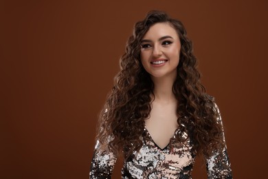 Photo of Beautiful young woman with long curly hair in sequin dress on brown background, space for text