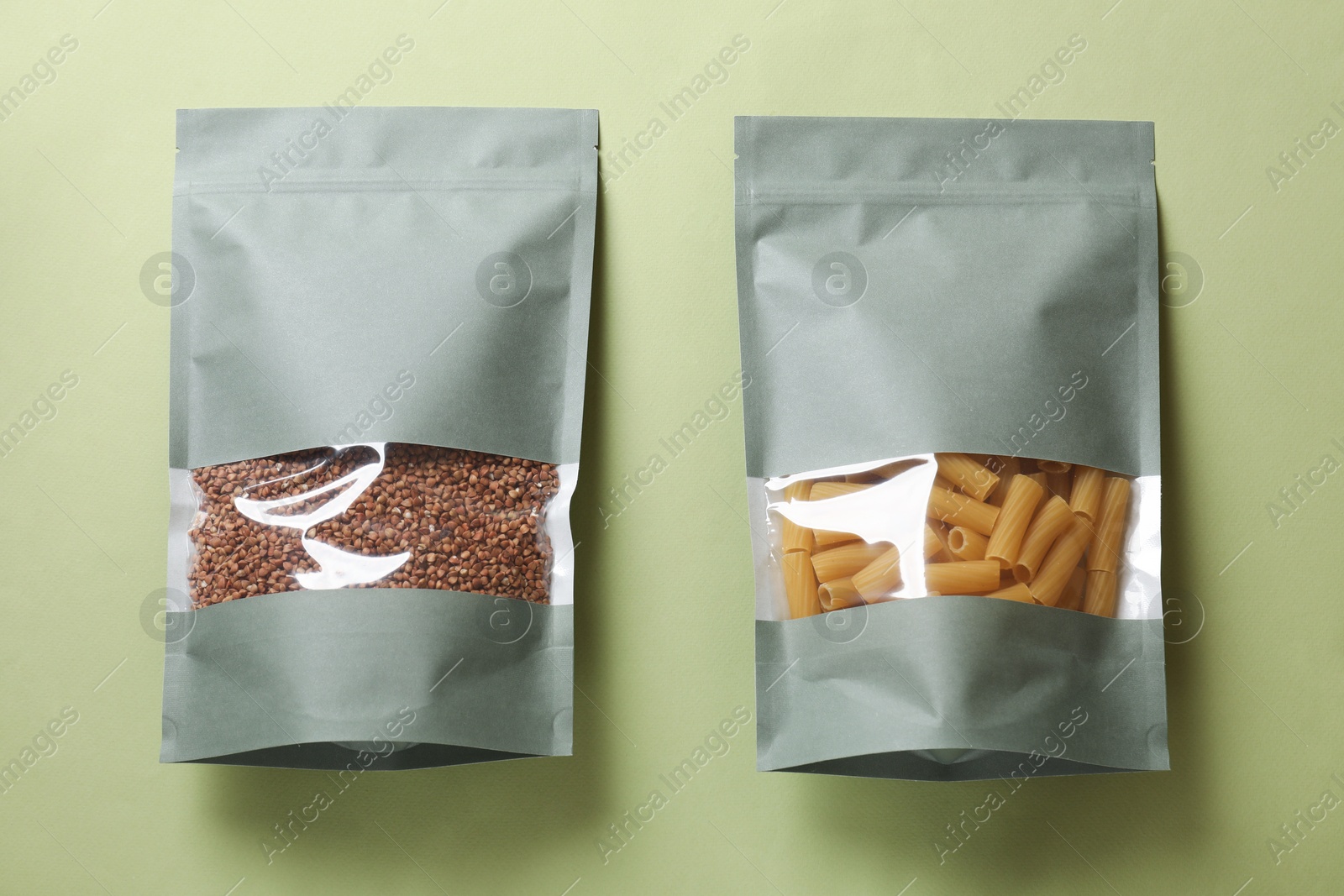 Photo of Paper pouch bags with buckwheat and pasta on green background, top view