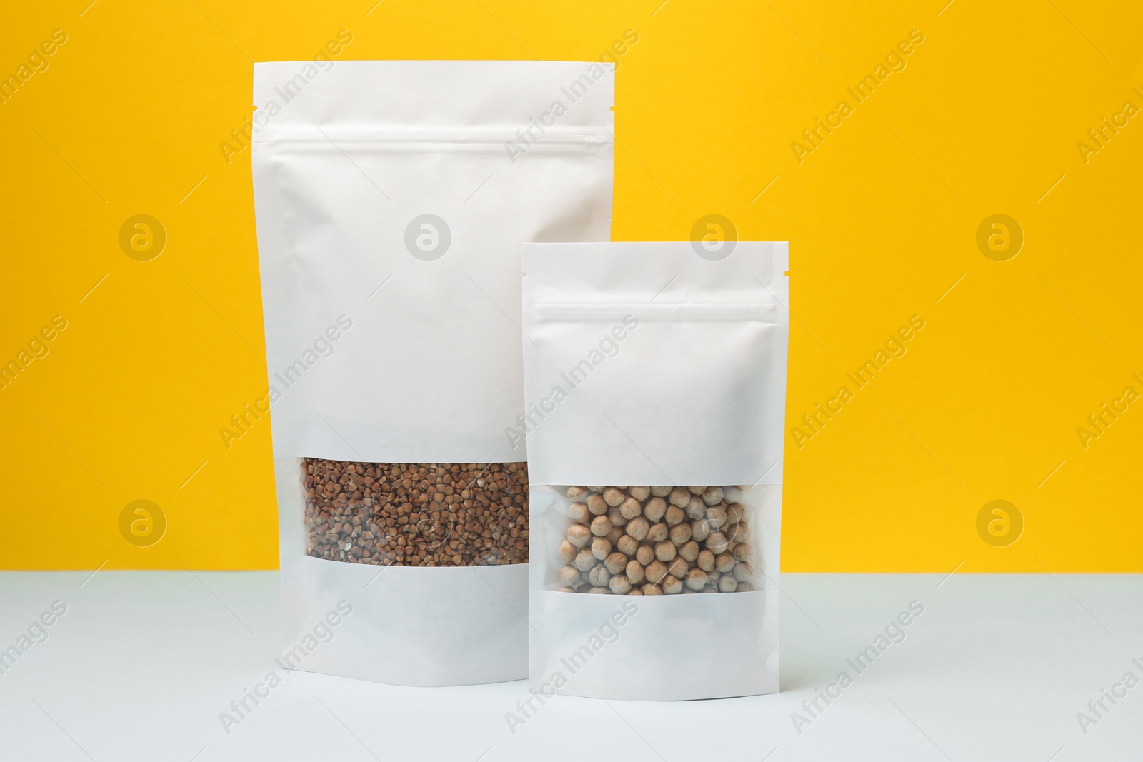 Photo of Paper pouch bags with chickpeas and buckwheat on table against yellow background