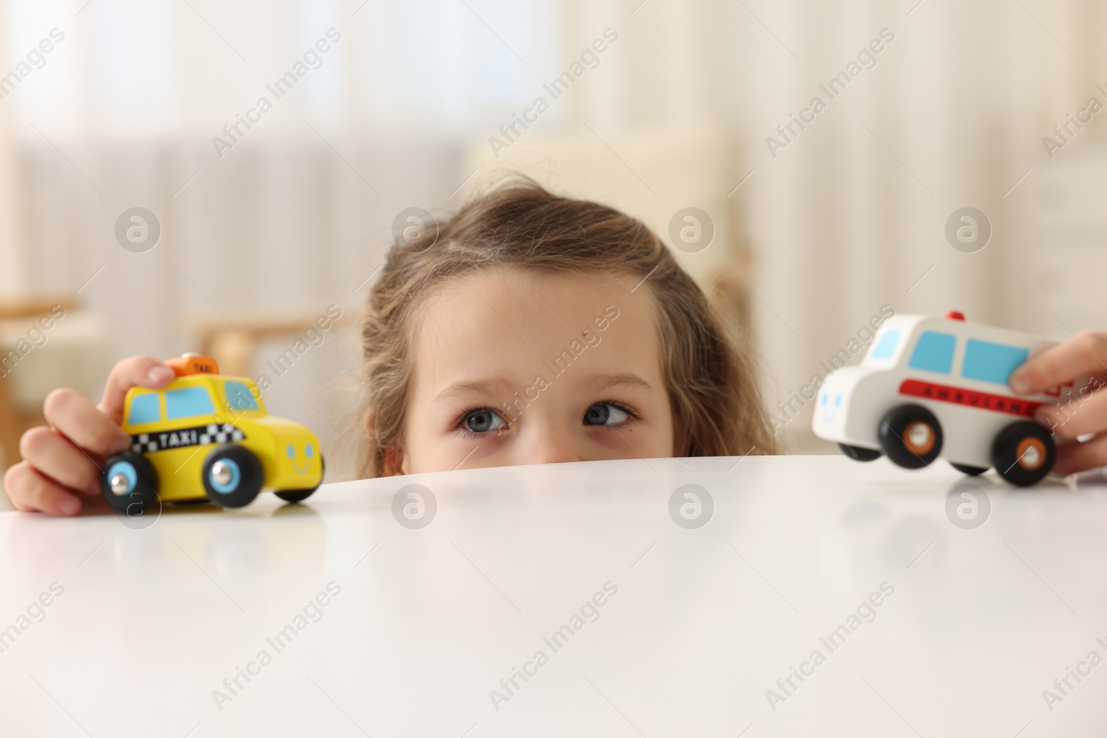 Photo of Little girl playing with toy cars at white table indoors, closeup