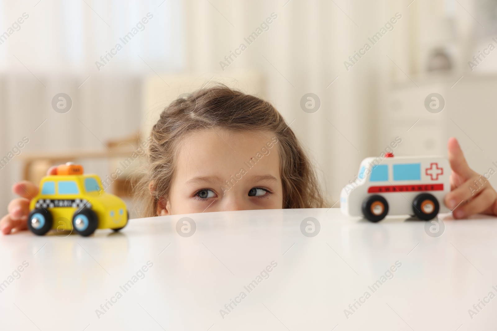 Photo of Little girl playing with toy cars at white table indoors, closeup