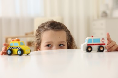 Photo of Little girl playing with toy cars at white table indoors, closeup