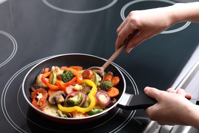 Photo of Woman cooking vegetables and mushrooms in frying pan on cooktop indoors, closeup