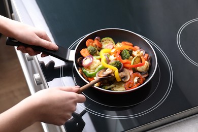 Photo of Woman cooking vegetables and mushrooms in frying pan on cooktop indoors, closeup