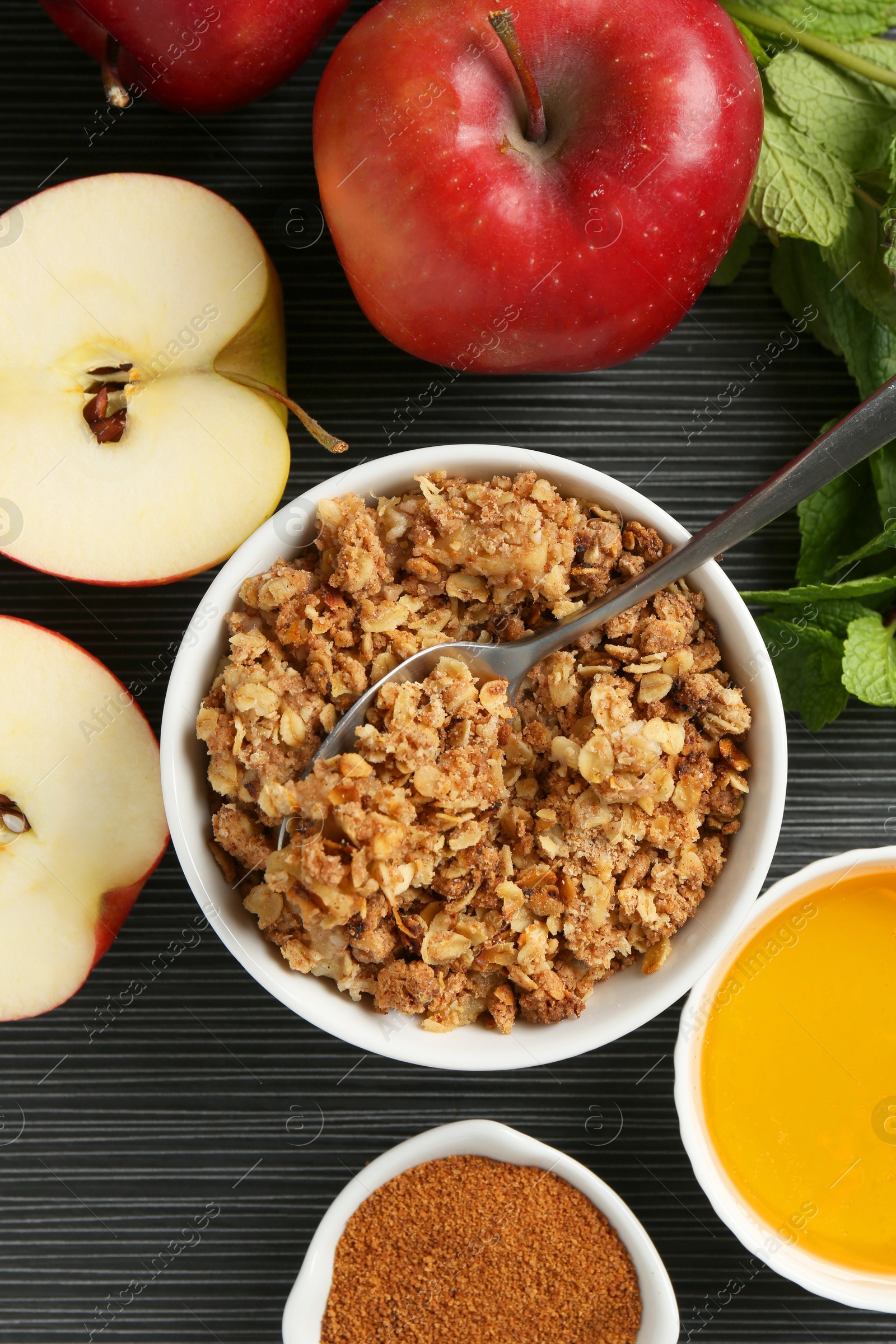 Photo of Delicious apple crisp in bowl, fresh fruits, cinnamon and mint on black textured table, flat lay